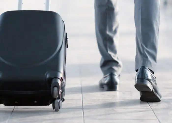 Man walking down a glass hallway in an airport pulling along a rolling carry-on suitcase