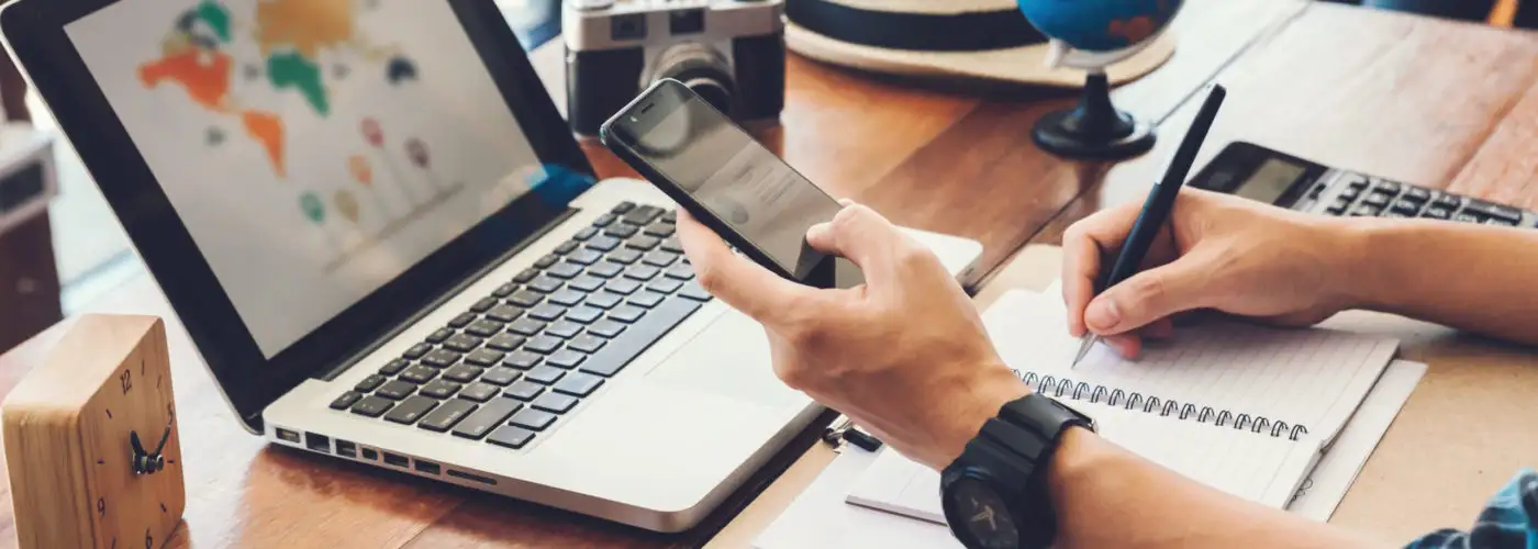 Close up of person writing in notebook, using phone and laptop to plan a trip