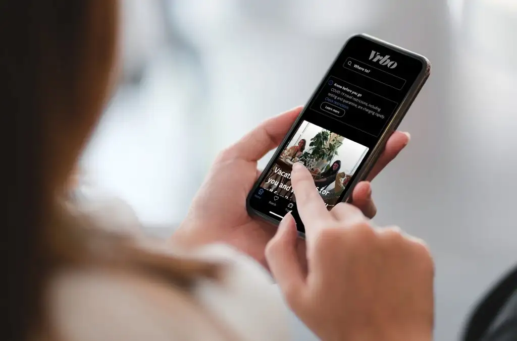 A man holding a phone showing the vacation rental bookingapp VRBO