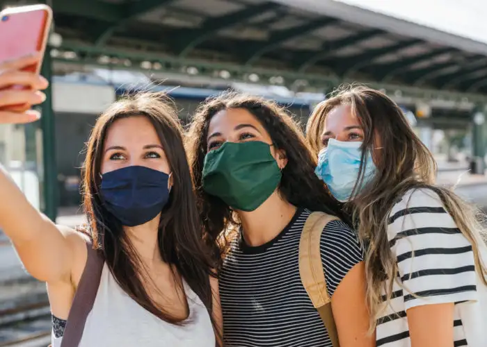 Three young friends taking a selfie in a train station wearing face masks