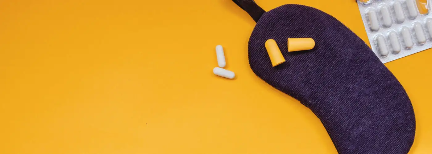 Sleep mask, eargplugs, and sleeping pills on yellow background