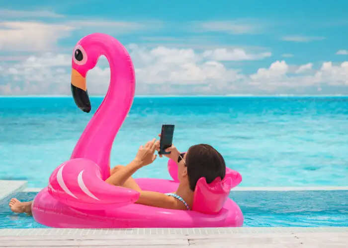 Woman lounging on inflatable flamingo in pool overlooking the ocean scrolling on smartphone