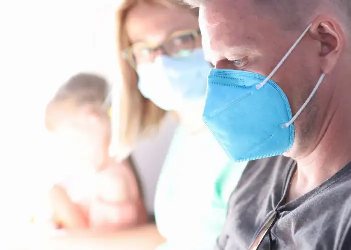 Man sits on airplane wearing a medical face mask with woman and child sitting next to him in the same row, out of focus