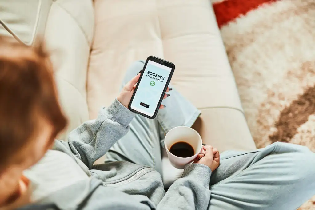 Woman booking flight on phone while drinking coffee on the couch