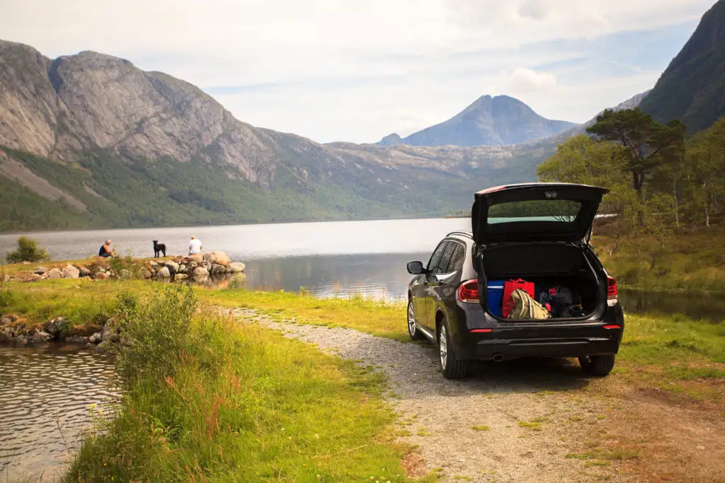 Car with trunk open pulled over to the side of a dirt road while the driver and passengers explore the shore of a lake in the distance