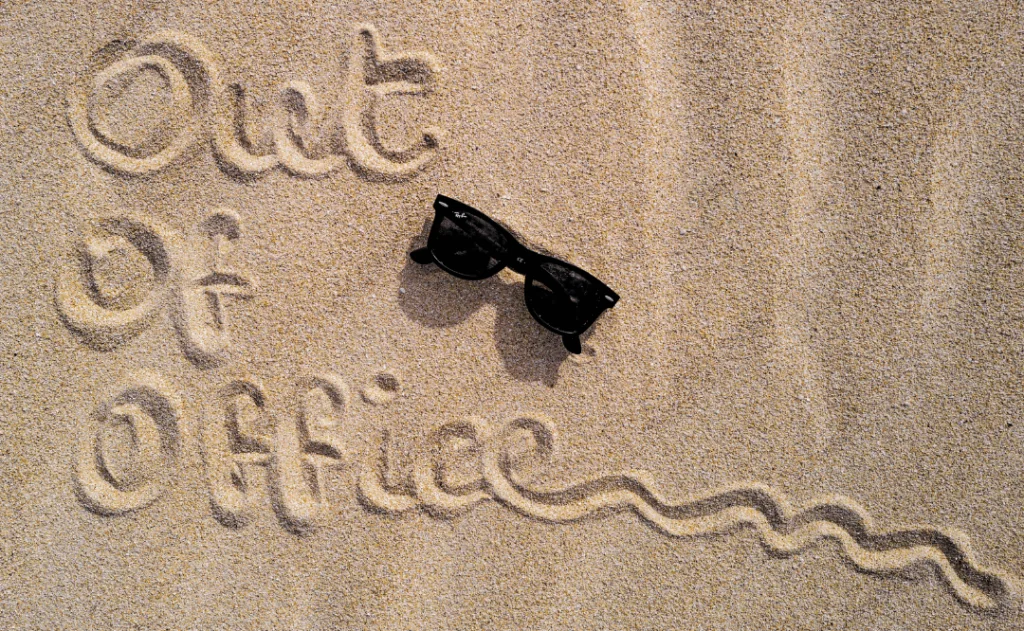 Out of office scribbled in the sand with RayBan sun glasses next to it