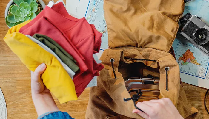 Person packing up a backpack for a trip surrounded by maps, a camera, and other travel gear