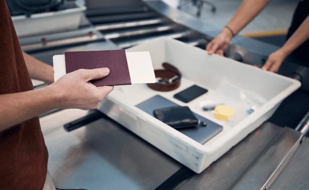 Passenger holding passport against personal Items, liquids, and laptop in container at airport security check..
