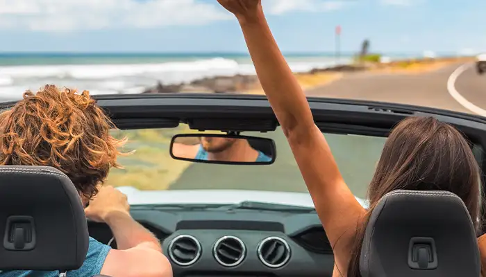 Road trip summer vacation fun young couple driving convertible car on holiday travel destination banner panoramic. Woman with arms up happy, man driver having fun.